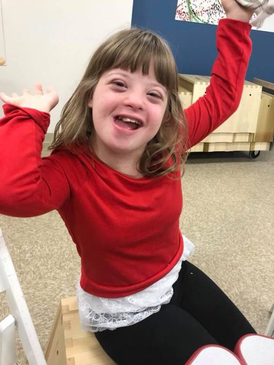 Little girl with Down syndrome wearing a red sweater and pumping her arms up.