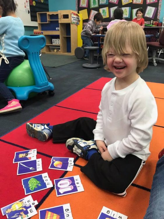 Little boy with Down syndrome at school, sitting on a rug, flashcards on the floor and he smiles at camera.