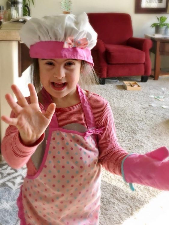 Little girl with Down syndrome wearing a pink chef's costume, including hat, apron and over mitt. She is waving at the camera with her other hand.