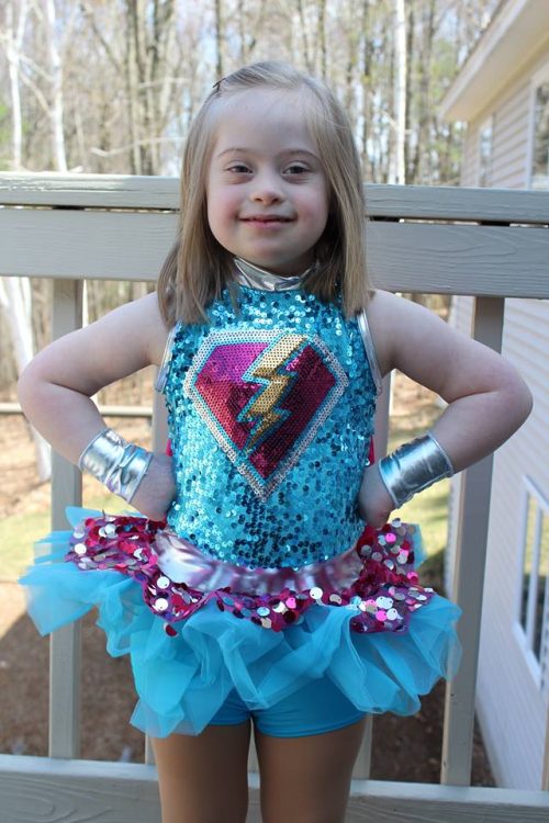 Little girl with Down syndrome wearing an unknown super hero outfit (blue with a purple diamond and a yellow flash symbol) and posing with her hands on her hips. 