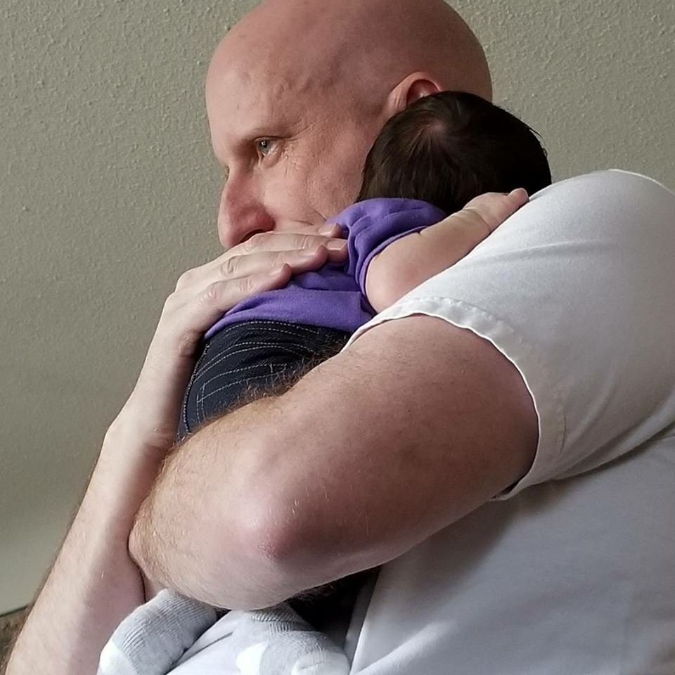 man in a white shirt holding his baby granddaughter against his chest