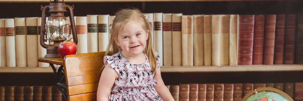 Little girl with down syndrome sitting on small wooden bench with a bookshelf as background.