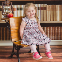 Little girl with down syndrome sitting on small wooden bench with a bookshelf as background.