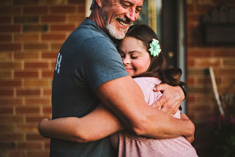 father hugging daughter with Down syndrome, daughter is closing her eyes, enjoying the hug, dad is happy and smiling. Daughter seems to be a teenager or young adult.