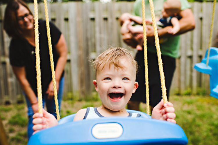 Boy with Down syndrome in blue swing, mom and dad and baby sibling blurry in the background