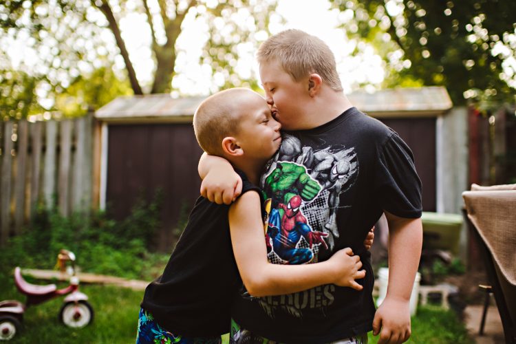 Two brothers hugging. Older brother has Down syndrome and kissing younger brother on forehead. Little brother is closing his eyes, letting the love in, and hugging his brother tight.