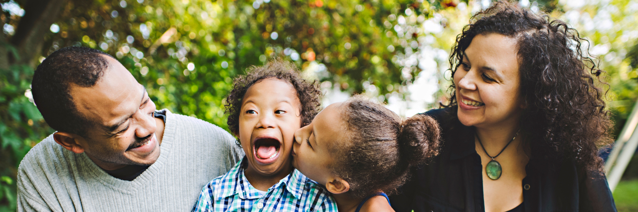 'Better With You' Highlights Families of Kids With Down Syndrome Family of four, mom and dad at the ends, sister kissing brother with Down syndrome who is smiling big and happy