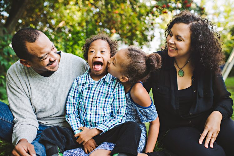 Family of four, mom and dad at the ends, sister kissing brother with Down syndrome who is smiling big and happy