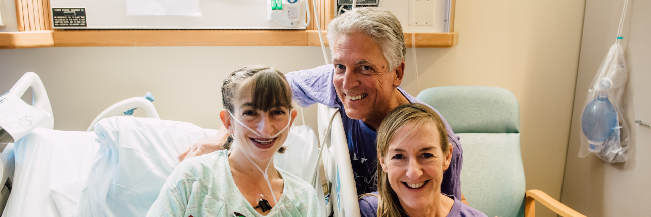 Parents of Kids With Disabilities, Raise Survivors, Not Victims Girl sitting in hospital bed with oxygen and monitors, she is smiling, posing with her dad and mom.