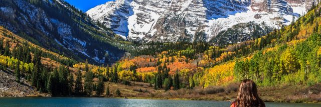 A woman admiring the autumnal beauty of the snowcapped Maroon Bells and the colorful foliage of the aspen groves on a sunny, cloudless day.