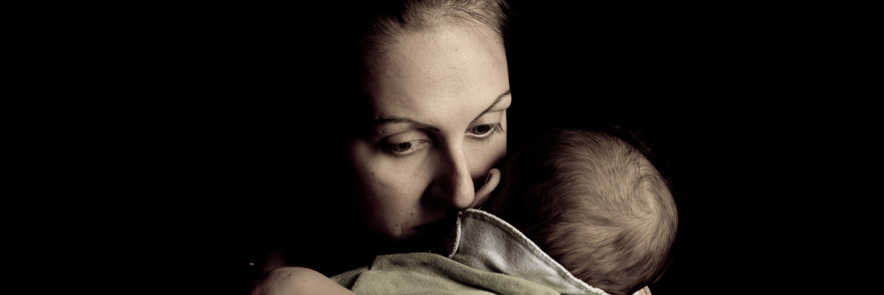 Black shadowed background, focus on mother hugging her baby and looking sad.