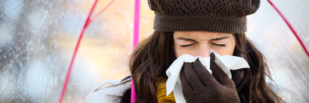 Why I Fear the Cold and Flu Season A woman walking outside with her umbrella, sneezing into a tissue.