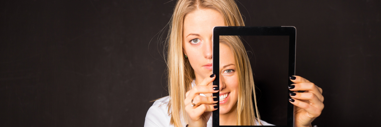 What My Smile Has Been Hiding Since My Chronic Pain Diagnosis A woman moving a tablet (showing a picture of her smiling), away from her face to show a serious expression.