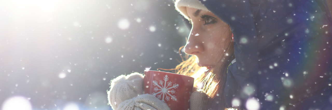 Living Through the Cold of Winter With Chronic Pain woman standing outside in the snow wearing a hooded jacket and gloves and holding a red mug