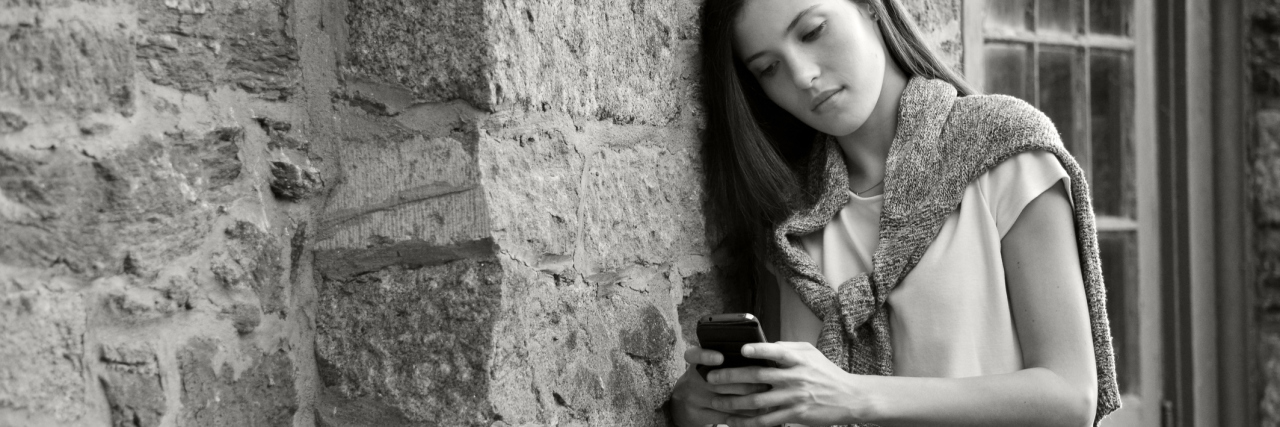 black and white photo of young woman on smartphone social media leaning against stone wall