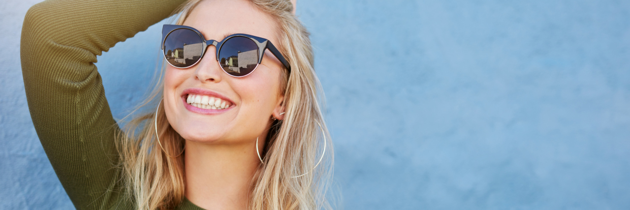 woman with blonde hair smiling and wearing sunglasses against a blue wall