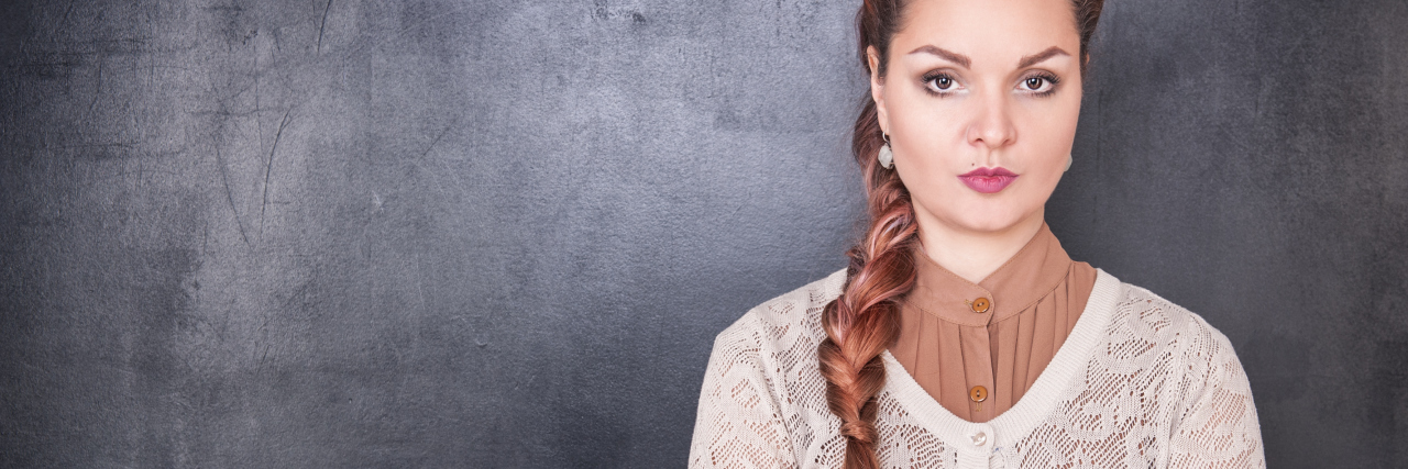 How Attitudes Toward Me Changed After I Received a Diagnosis woman with long hair in a braid standing against a wall with her arms crossed