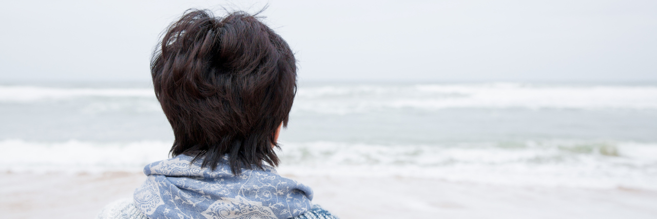 Why It's Harmful to Be in Denial About Chronic Illness woman standing on the beach and wrapped in a blue knit blanket