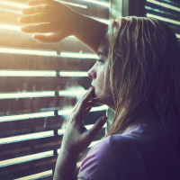 An upset woman looking through the blinds, letting some sunlight in the room.