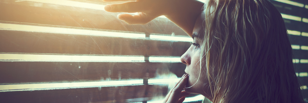 Sad and lonely blonde woman with wet hair looking through window blinds into the sunlight