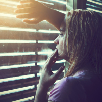 Sad and lonely blonde woman with wet hair looking through window blinds into the sunlight