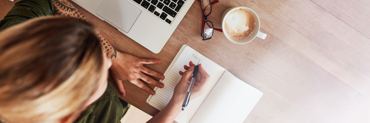 New Year's Resolutions as a Person with Chronic Illness woman writing and sitting at a table with her laptop and coffee