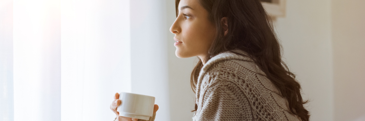 dark haired woman looking out window with coffee