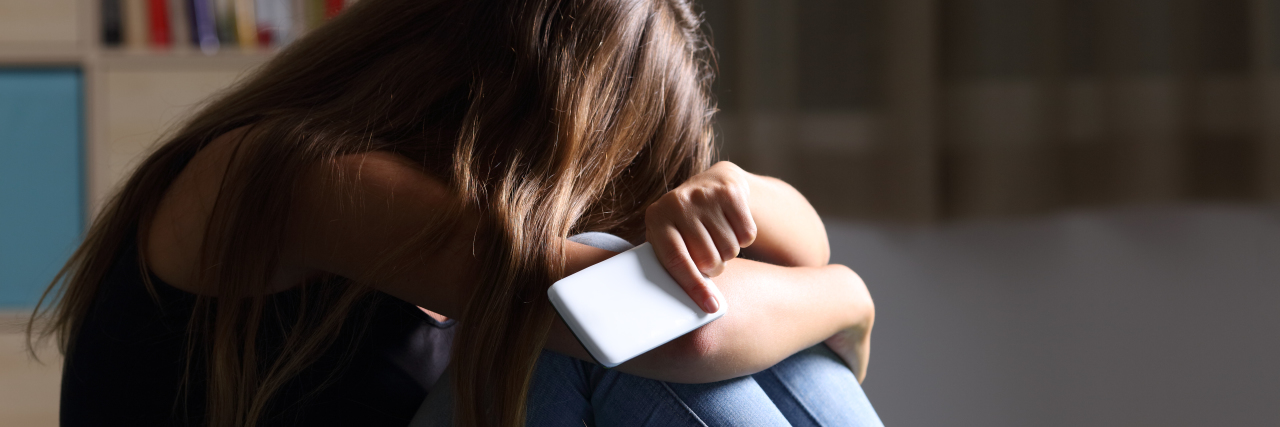 Anxiety of Telling Someone About Your Borderline Personality Disorder Single sad teen holding a mobile phone lamenting sitting on the bed in her bedroom with a dark light in the background