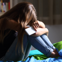 Single sad teen holding a mobile phone lamenting sitting on the bed in her bedroom with a dark light in the background