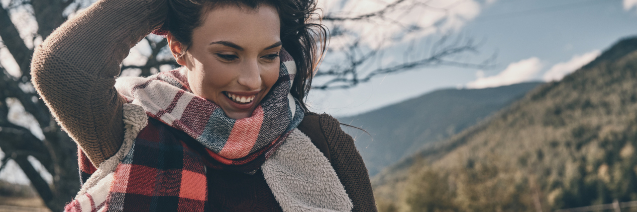 Embracing My Imperfections as a Woman With Chronic Illness woman wearing a scarf and jacket standing outside smiling and running her fingers through her hair