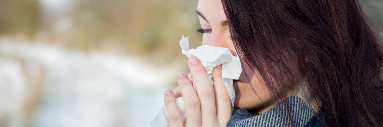 The Difference in Having the Cold or Flu Versus a Chronic Illness woman wearing a blue scarf and blowing her nose outside
