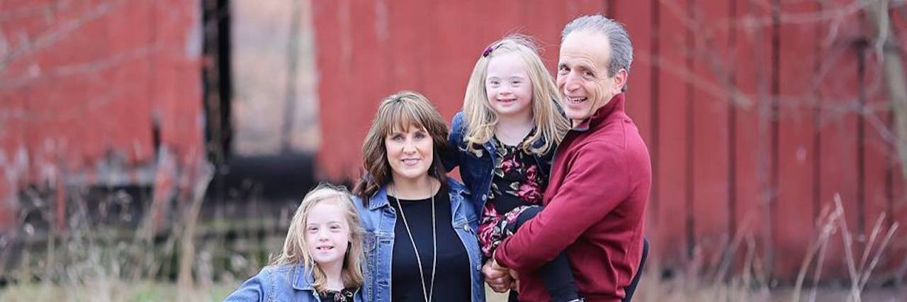 My Child With Down Syndrome Is More Than a Girl With Pink Headphones Family of four, mom and dad and two girls with Down syndrome, posing outdoors with a red barn in the background