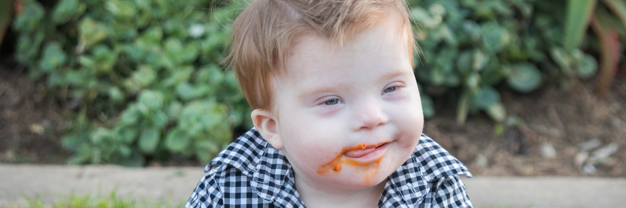 Letting Go of Typical Expectations for My Son With Down Syndrome Little boy with Down syndrome on his first birthday sitting outside on blanket eating a cake with orange frosting and smiley face (maybe a crab cake)