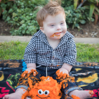 Little boy with Down syndrome on his first birthday sitting outside on blanket eating a cake with orange frosting and smiley face (maybe a crab cake)