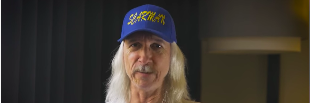 Image is Scarman wearing a Scarman blue baseball hat and white Scarman shirt