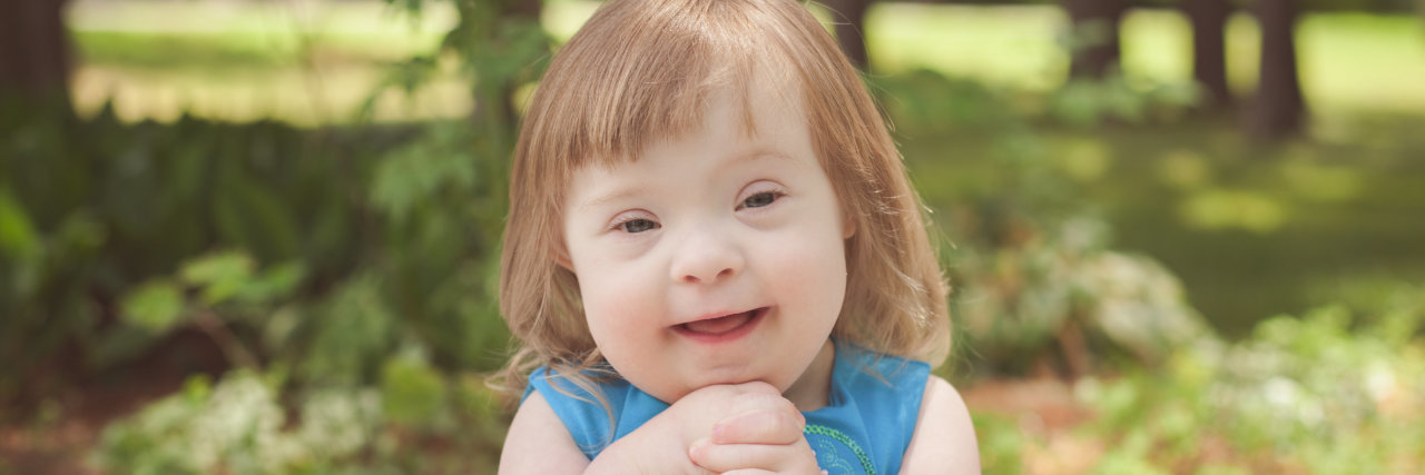 I Used to Think the R-Word Was Funny Little girl with Down syndrome wearing a blue dress and holding her hands together, smiling at camera.