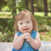 Little girl with Down syndrome wearing a blue dress and holding her hands together, smiling at camera.