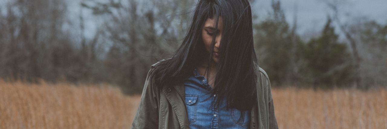 Being Jealous of Other People's Accomplishments With Chronic Illness woman in a jean shirt standing outside in a field with cloudy weather looking down