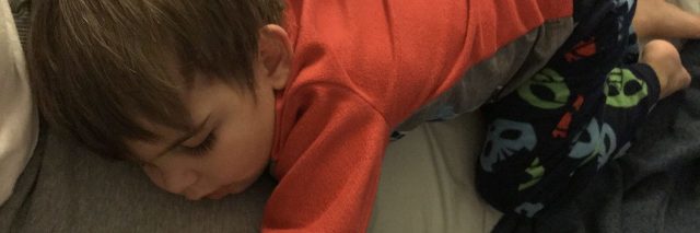 Little boy sleeping n top of his mom, he is wearing orange pajamas and mom has gray sweatpants on