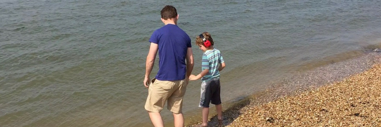 Father and son at the beach by the water. dad is wearng a royal blue shirt and holds his son's hand. Son is wearing red noise cancelling headphones and a striped sky blue shirt.
