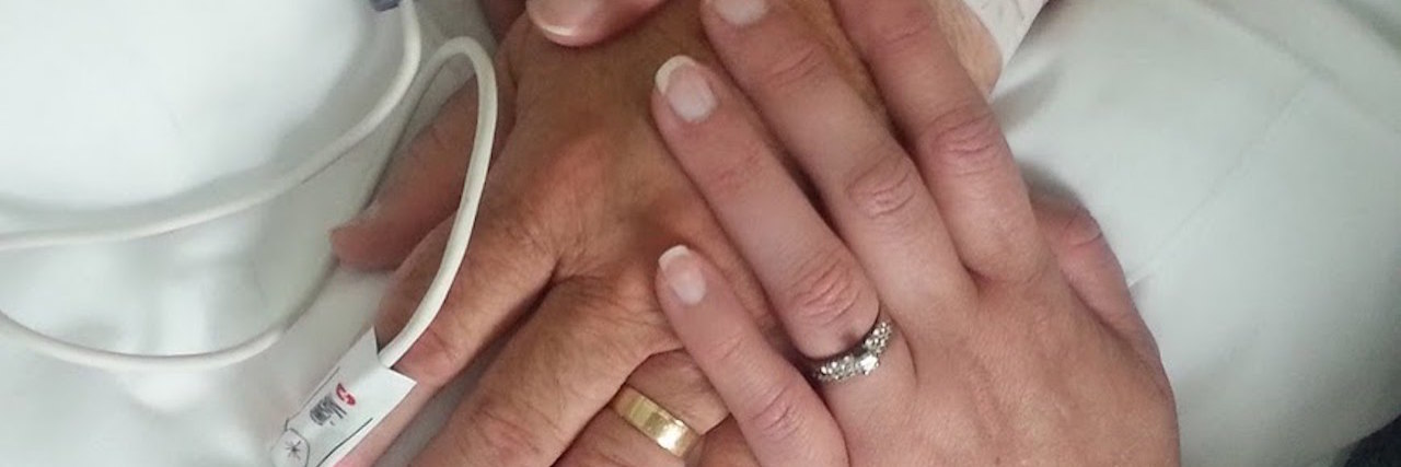 To Those Who Care for Someone With Chronic Illness wife holding husband's hand while he lies in a hospital bed connected to monitors