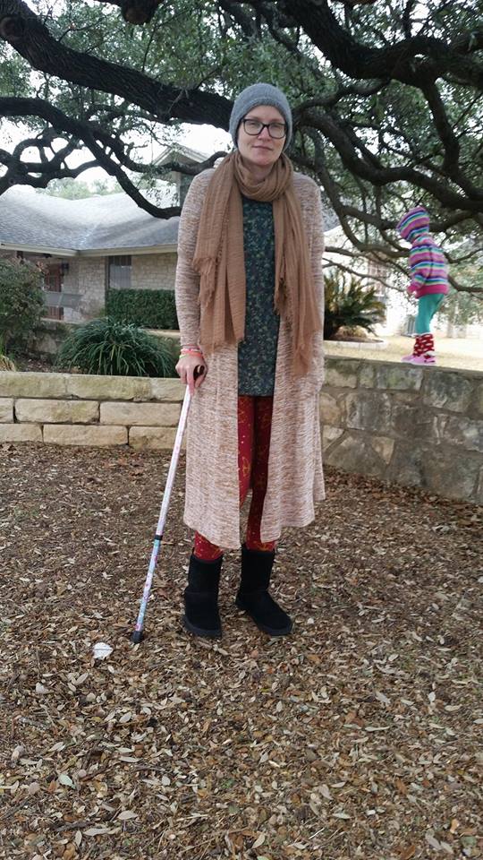 woman wearing several jackets and a robe standing outside with a cane