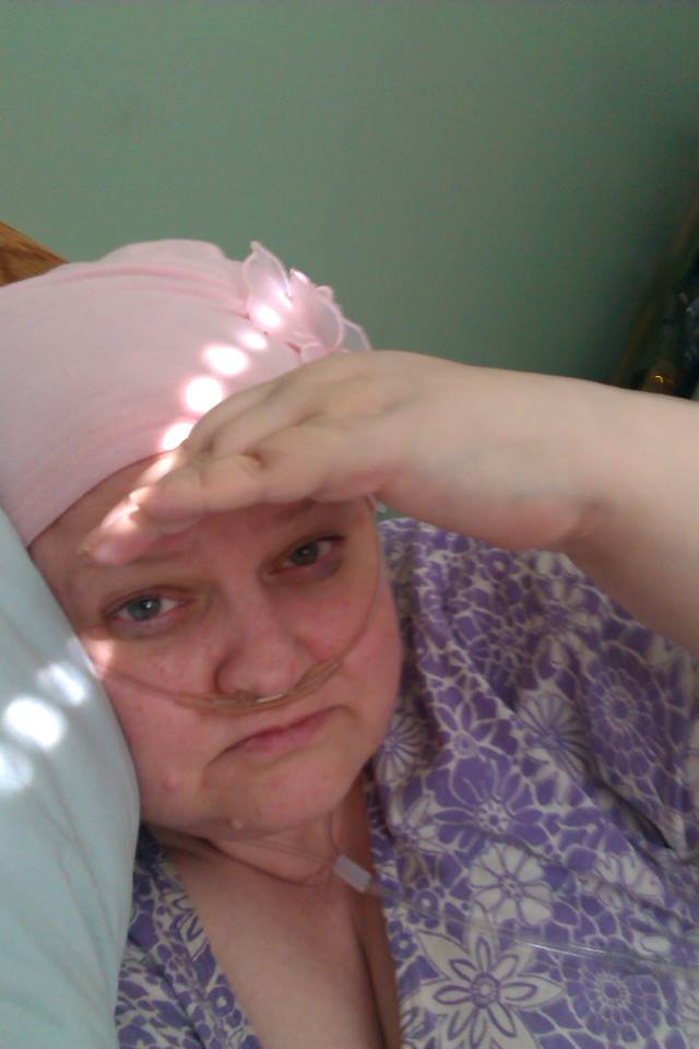 woman lying in bed with an oxygen tube in her nose and a pink scarf over her head