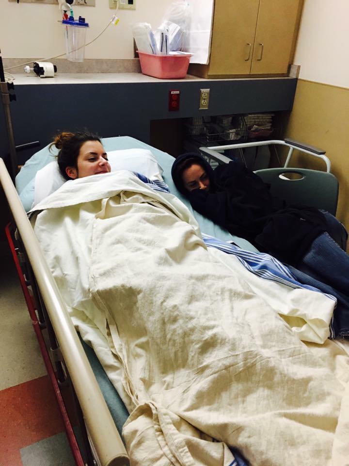 woman lying in a hospital bed covered with a blanket with her sister sitting next to her