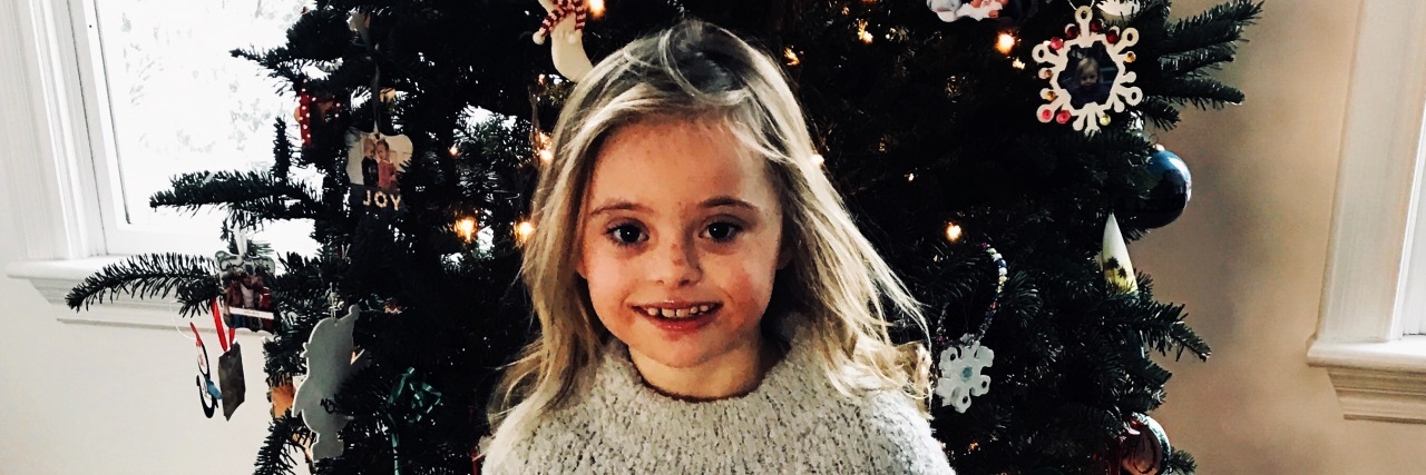 Little girl with Down syndrome standing in front of her Christmas tree and smiling for picture