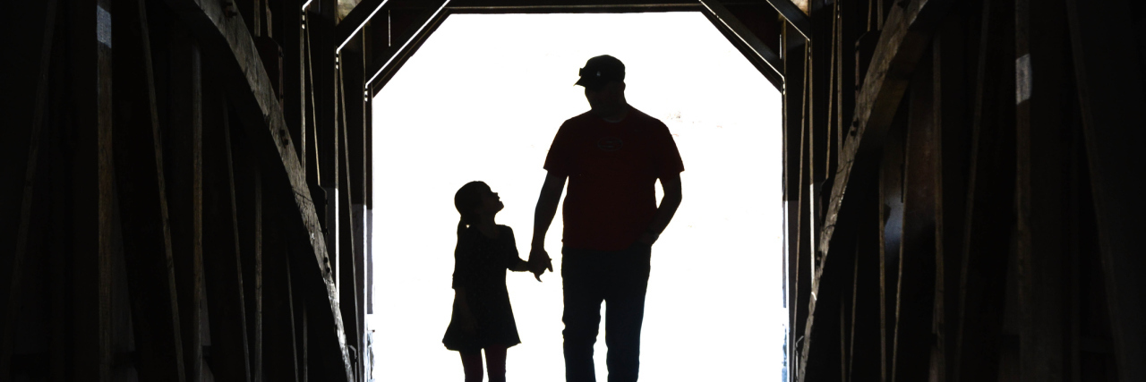 A silhouette image of a father and daughter holding hands as they walk down a tunnel.