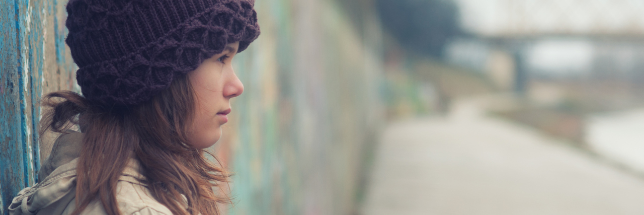 What It's Like When a Fibromyalgia Flare Begins young woman in a white jacket and purple beanie leaning against a wall outside on a cloudy day