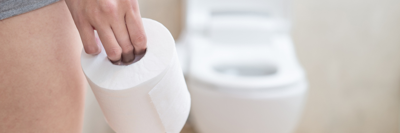 A woman standing in front of a toilet, holding toilet paper.