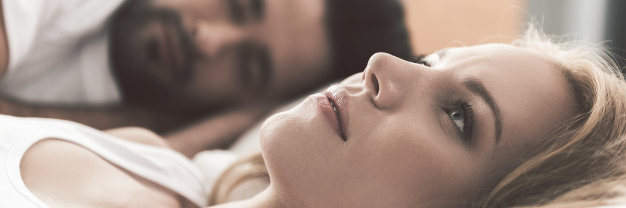 How Chronic Illness Can Affect a Relationship Nervous young woman is lying on bed and thinking seriously. Her husband is sleeping on background