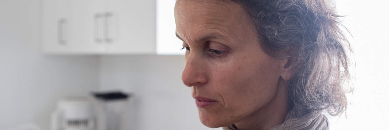 When Your Symptoms Aren't Your Chronic Illness A woman in her kitchen, staring off into space.